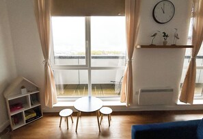 Ein Zimmer mit großem Fenster, einem runden Tisch mit vier Stühlen, einem blauen Sofa, einem Regal mit Dekoration und einer Wanduhr.