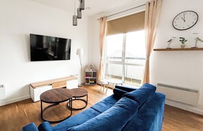 En stue med en blå sofa, et fjernsyn, et vindue med gardiner, et ur og et lille bord.
