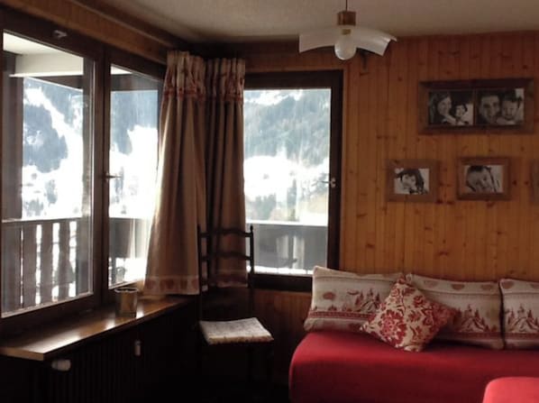 A cozy room with a wooden interior, a red sofa, a window with curtains, and framed pictures on the wall. - Beautiful Flat Facing La Clusaz Slopes (La Clusaz)