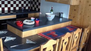 Una cocina compacta con microondas, salpicadero a cuadros, armarios de madera, una zona de comedor con mesa y sillas, y una botella de vino sobre la encimera.