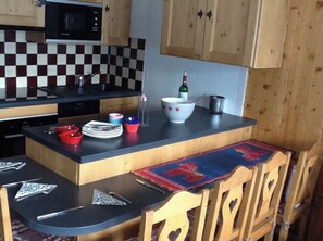 A compact kitchen with a microwave, checkered backsplash, wooden cabinets, a dining area with a table and chairs, and a bottle of wine on the counter. - Beautiful Flat Facing La Clusaz Slopes (La Clusaz)