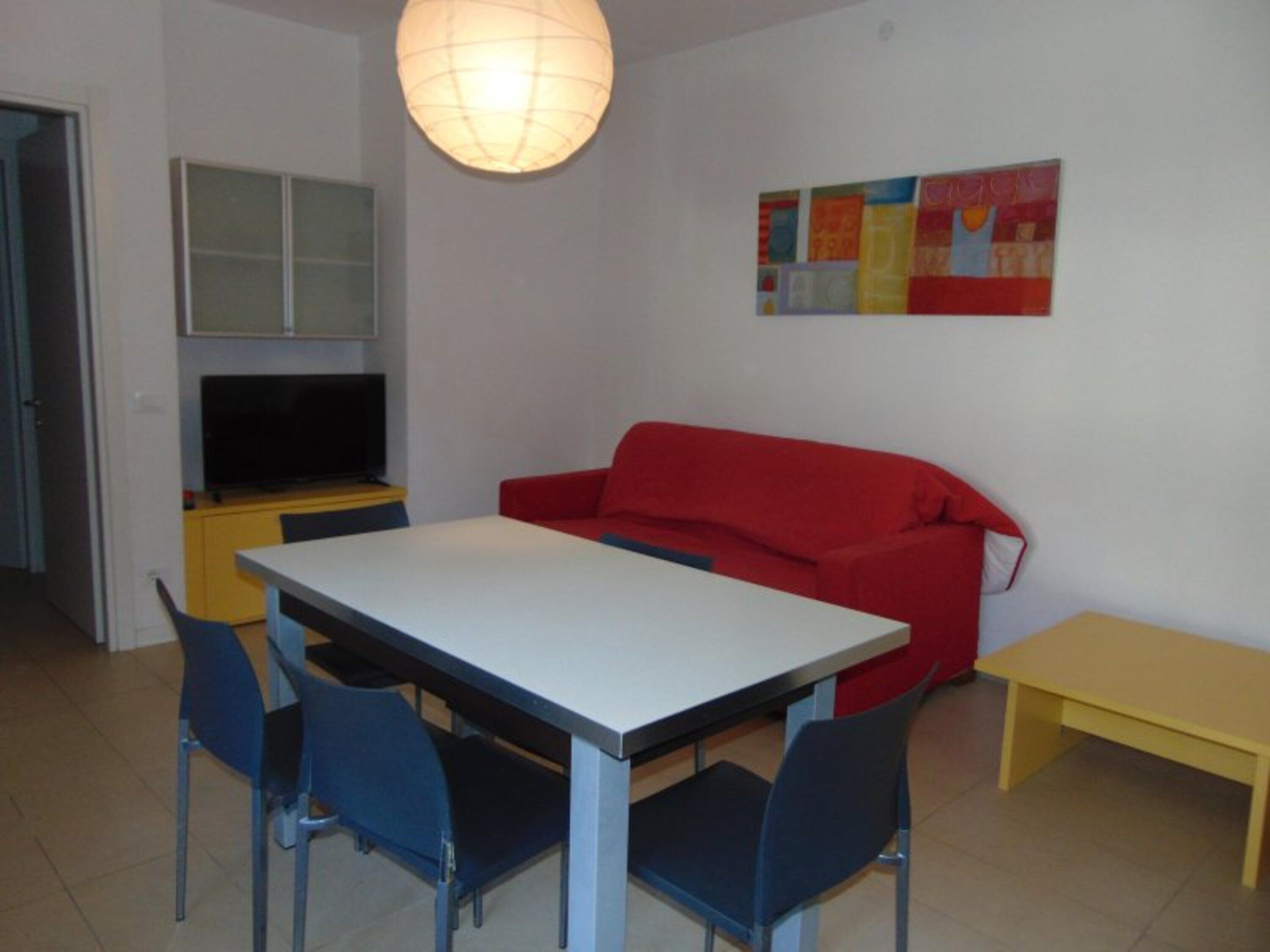 A small, modern living space with a dining table, chairs, a red sofa, a television, and a colorful artwork on the wall.