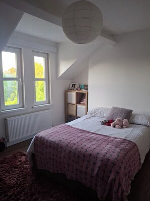 Een slaapkamer met een bed, een raam, een radiator, een plank met spullen en een plafondlamp.