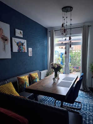 Een eetgedeelte met een houten tafel, een blauwe met stof beklede bank en uitzicht op de tuin door het raam.