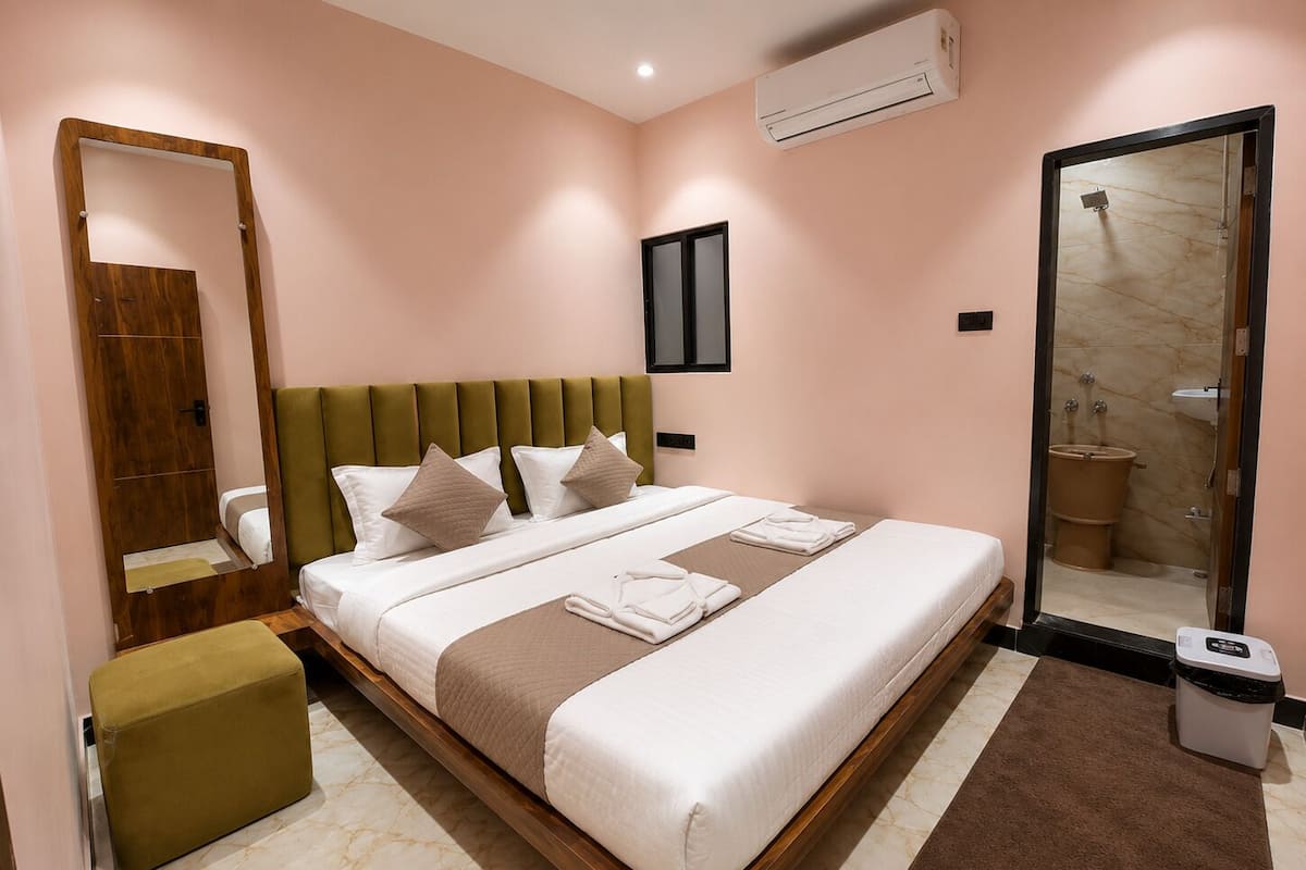 A hotel room with a bed, a mirror, a wardrobe, an air conditioning unit, and a bathroom visible through a glass door.