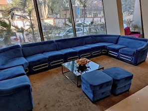 A modern living room with a blue sectional sofa, a glass coffee table, and a view of the street through large windows. - Ramaya The Boutique Hotel Haryana (Rohtak)