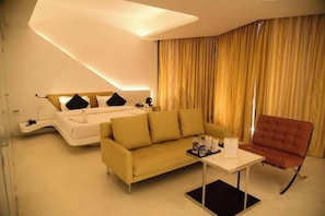 A modern hotel room with a sofa, a chair, a small table, and a bed with a headboard. - Ramaya The Boutique Hotel Haryana (Rohtak)