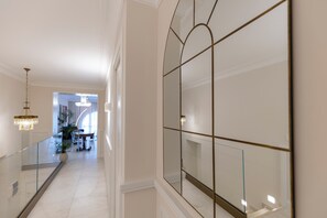 Un couloir doté d’une rambarde en verre, d’un lustre, d’un grand miroir et, au fond, d’un espace dédié à la salle à manger.