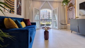Ein modernes Wohnzimmer mit einem blauen Sofa, einem Glastisch und Blick auf ein Wohngebäude durch große Fenster.