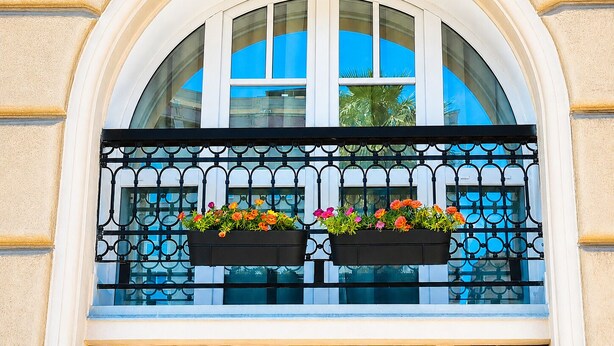 Ein rundbogenförmiges Fenster mit einem schwarzen schmiedeeisernen Balkon, zwei Blumenkästen und einem Palmspiegelbild.