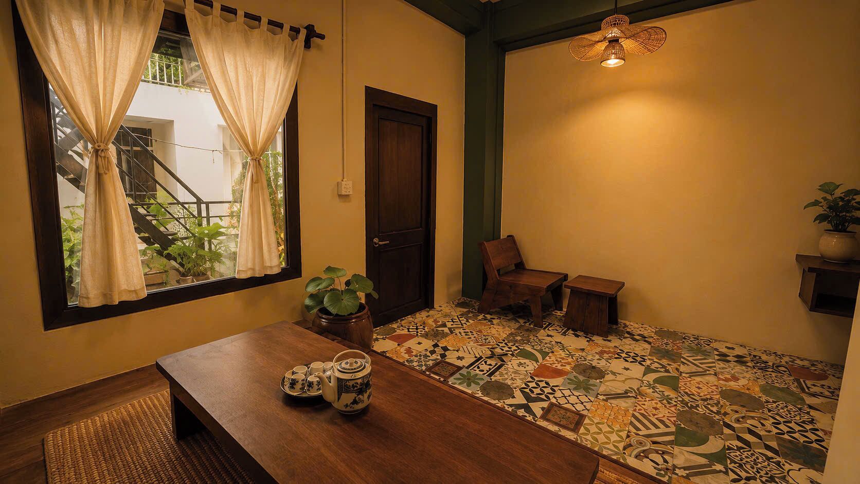 A room with a wooden table set with teacups and a teapot, a potted plant, a chair, a small table, a patterned floor, a door, and a hanging light fixture.