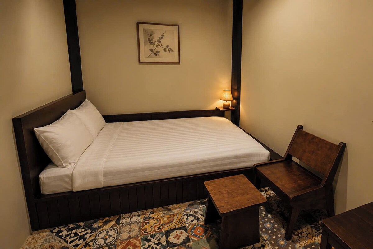 A neatly made bed with a pillow, a bedside table with a lamp, a chair, and a small table.