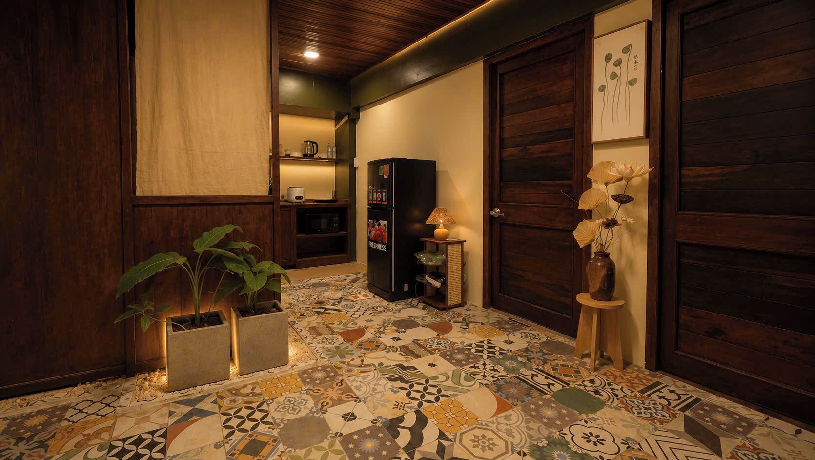 A hallway with a tiled floor, wooden doors, a refrigerator, a plant, and a small table with a lamp.