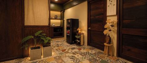 A hallway with a tiled floor, wooden doors, a refrigerator, a plant, and a small table with a lamp.