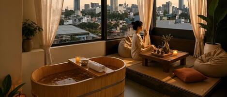 A spa room with a wooden bathtub, a view of a cityscape, and a person relaxing by the window.