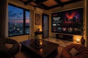 A cozy living room with a large window overlooking a cityscape, a flat-screen TV displaying a Netflix series, and comfortable seating. - Nha Cuoi Boutique Serviced Aparthotel (Ho Chi Minh City)