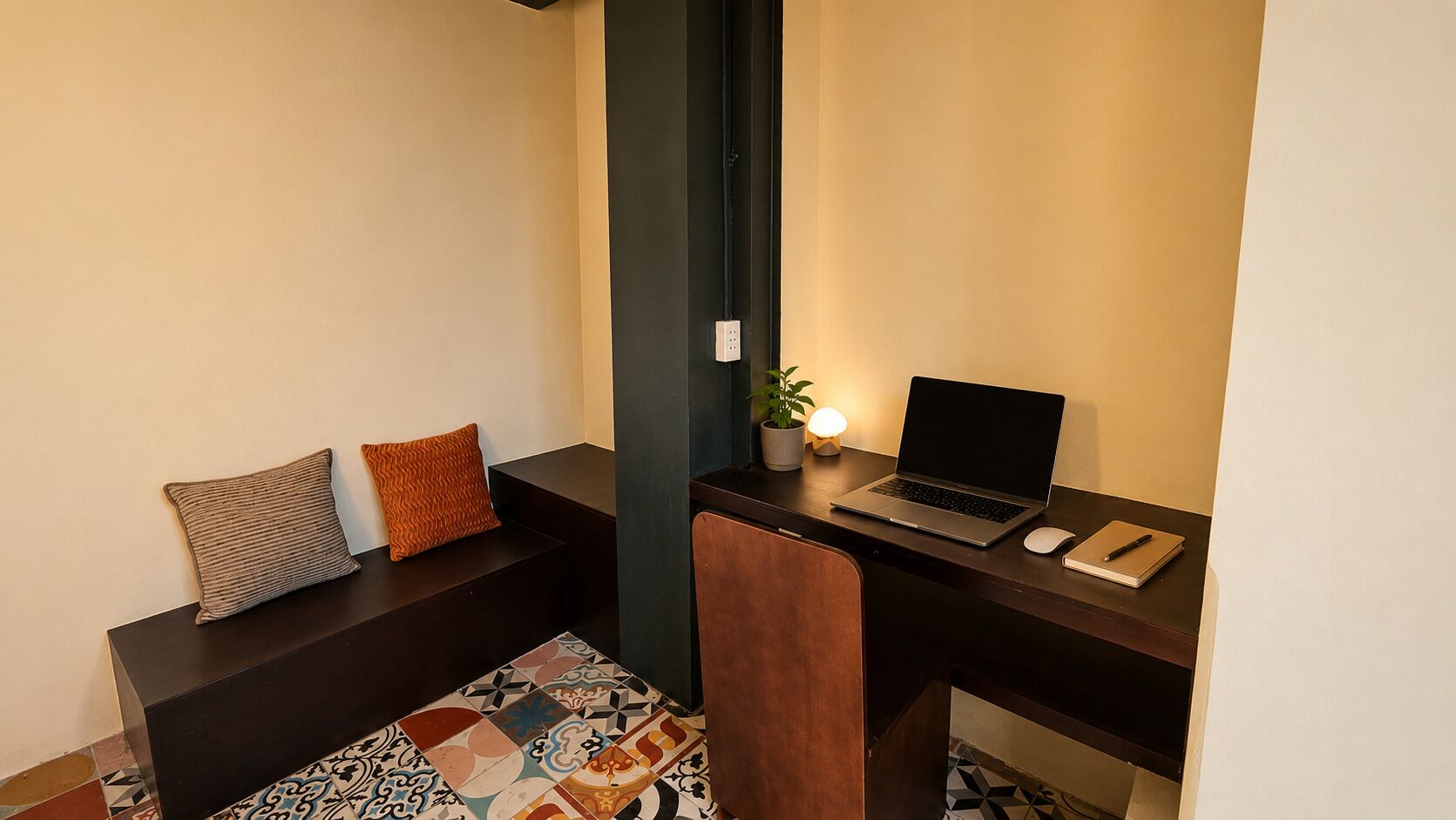 A corner room with a desk, laptop, chair, two pillows, a small plant, and a patterned rug.