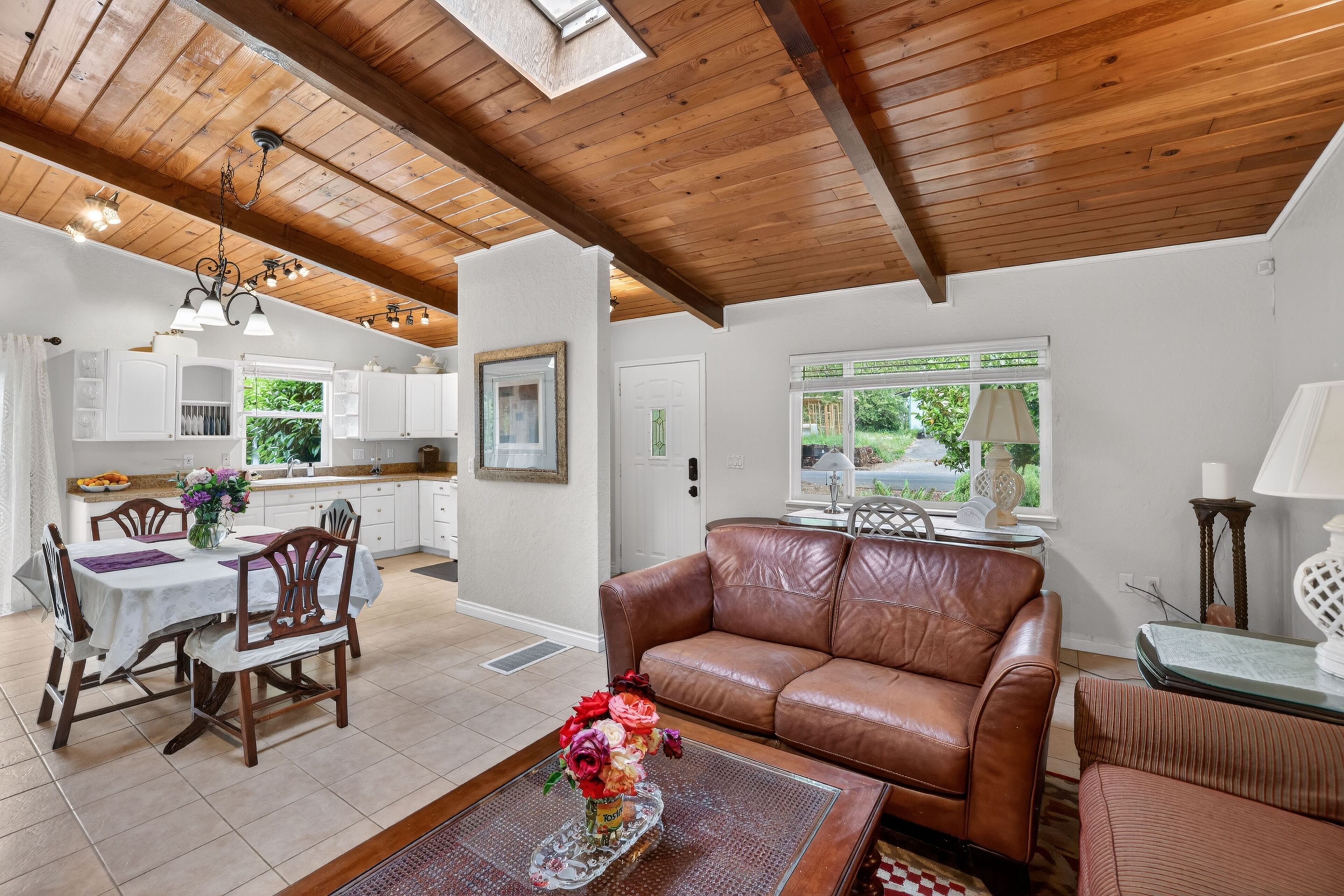 A spacious living area with a wooden ceiling, a dining table with chairs, a brown leather sofa, and a window with a view of greenery.