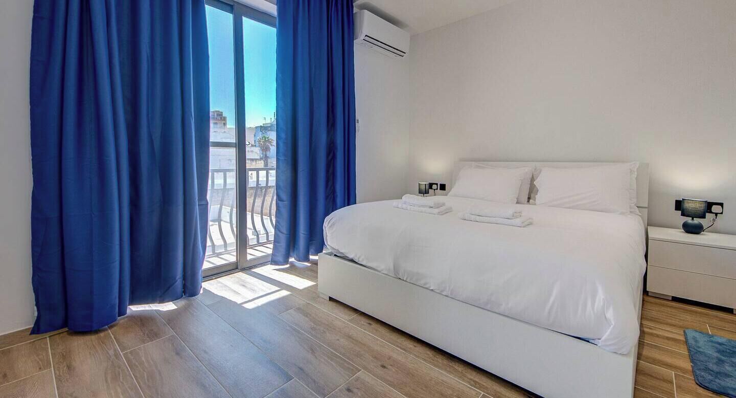 Een slaapkamer met een bed, een nachtkastje, een lamp en uitzicht door een raam met gordijnen.