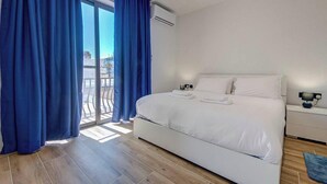 A bedroom with a bed, a nightstand, a lamp, and a view through a window with curtains. - 2BR Maisonette Near St. Paul`s Bay & Beaches (St. Paul`s Bay)