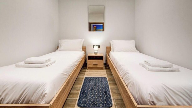 Quarto duplo com duas camas, uma mesinha de cabeceira, um abajur, um tapete pequeno e uma pintura na parede.