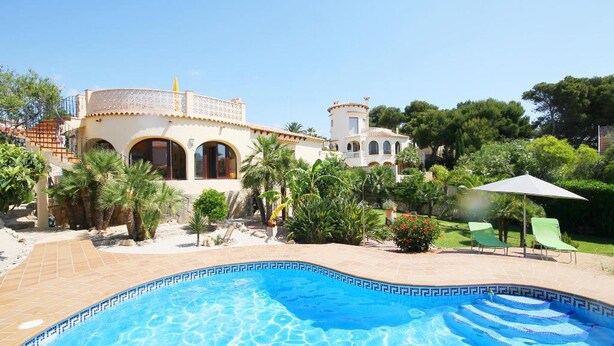 A pool with clear blue water, surrounded by a garden with palm trees and a building with a tiled roof. - Villa- Calypso in J vea (Javea)