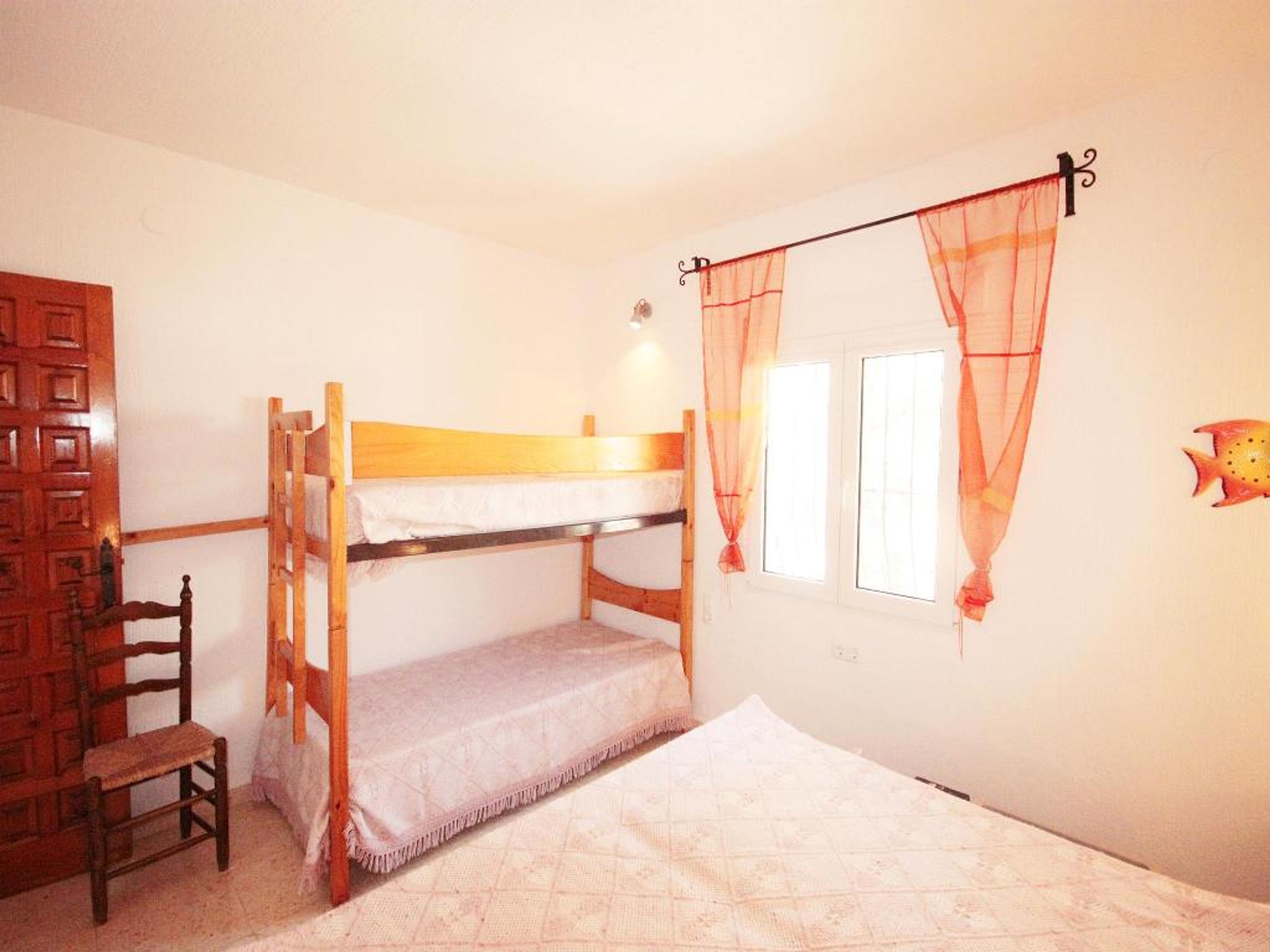 Habitación con literas, una silla de madera, una ventana con cortinas translúcidas y una decoración en la pared.