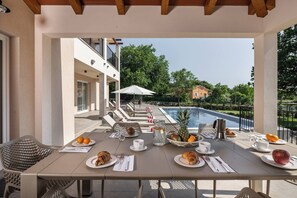 Ein Essbereich im Freien mit einem für das Frühstück gedeckten Tisch, mit Blick auf einen Swimmingpool und einen Garten.