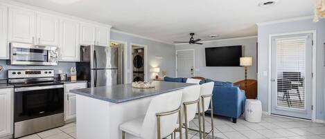 Una cocina moderna con armarios blancos, electrodomésticos de acero inoxidable, una isla central con taburetes altos, un televisor de pantalla plana y un ventilador de techo.