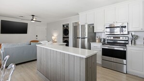 A modern kitchen with white cabinets, stainless steel appliances, and a grey sofa in the living area. - Coastal 4BR/4BA coastal duplex featuring king and queen ensuite bedrooms, a fully stocked kitchen, fenced pet-friendly yard, outdoor shower, beach gear, Smart TVs, WiFi, and parking for up to 6+ vehicles. (Wrightsville Beach)