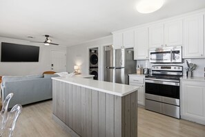 A modern kitchen with white cabinets, stainless steel appliances, and a grey sofa in the living area. - Coastal 4BR/4BA coastal duplex featuring king and queen ensuite bedrooms, a fully stocked kitchen, fenced pet-friendly yard, outdoor shower, beach gear, Smart TVs, WiFi, and parking for up to 6+ vehicles. (Wrightsville Beach)