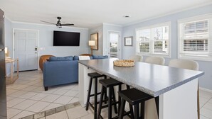 A modern kitchen with a central island, bar stools, a blue sofa, and a ceiling fan. - Coastal 4BR/4BA coastal duplex featuring king and queen ensuite bedrooms, a fully stocked kitchen, fenced pet-friendly yard, outdoor shower, beach gear, Smart TVs, WiFi, and parking for up to 6+ vehicles. (Wrightsville Beach)