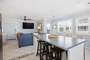 A modern kitchen with a central island, bar stools, a blue sofa, and a ceiling fan. - Coastal 4BR/4BA coastal duplex featuring king and queen ensuite bedrooms, a fully stocked kitchen, fenced pet-friendly yard, outdoor shower, beach gear, Smart TVs, WiFi, and parking for up to 6+ vehicles. (Wrightsville Beach)