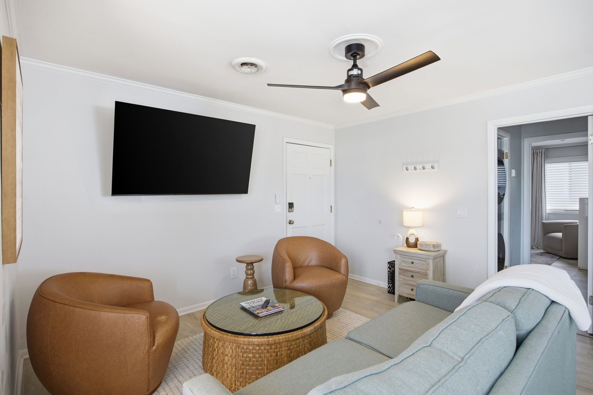 A living room with a flat-screen TV, two leather armchairs, a round coffee table, a sofa, a ceiling fan, and a small bedside table with a lamp.