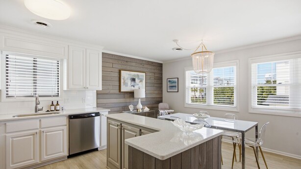 A modern kitchen with white cabinetry, a wooden accent wall, a central island, and a dining area with clear chairs. - Coastal 4BR/4BA coastal duplex featuring king and queen ensuite bedrooms, a fully stocked kitchen, fenced pet-friendly yard, outdoor shower, beach gear, Smart TVs, WiFi, and parking for up to 6+ vehicles. (Wrightsville Beach)