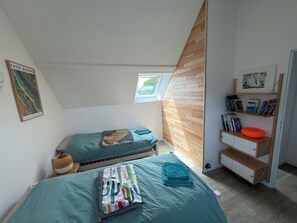 Un dormitorio con techo inclinado, una ventana, una cama con manta temática de cactus, una mesita de noche y una estantería con libros y un objeto decorativo.