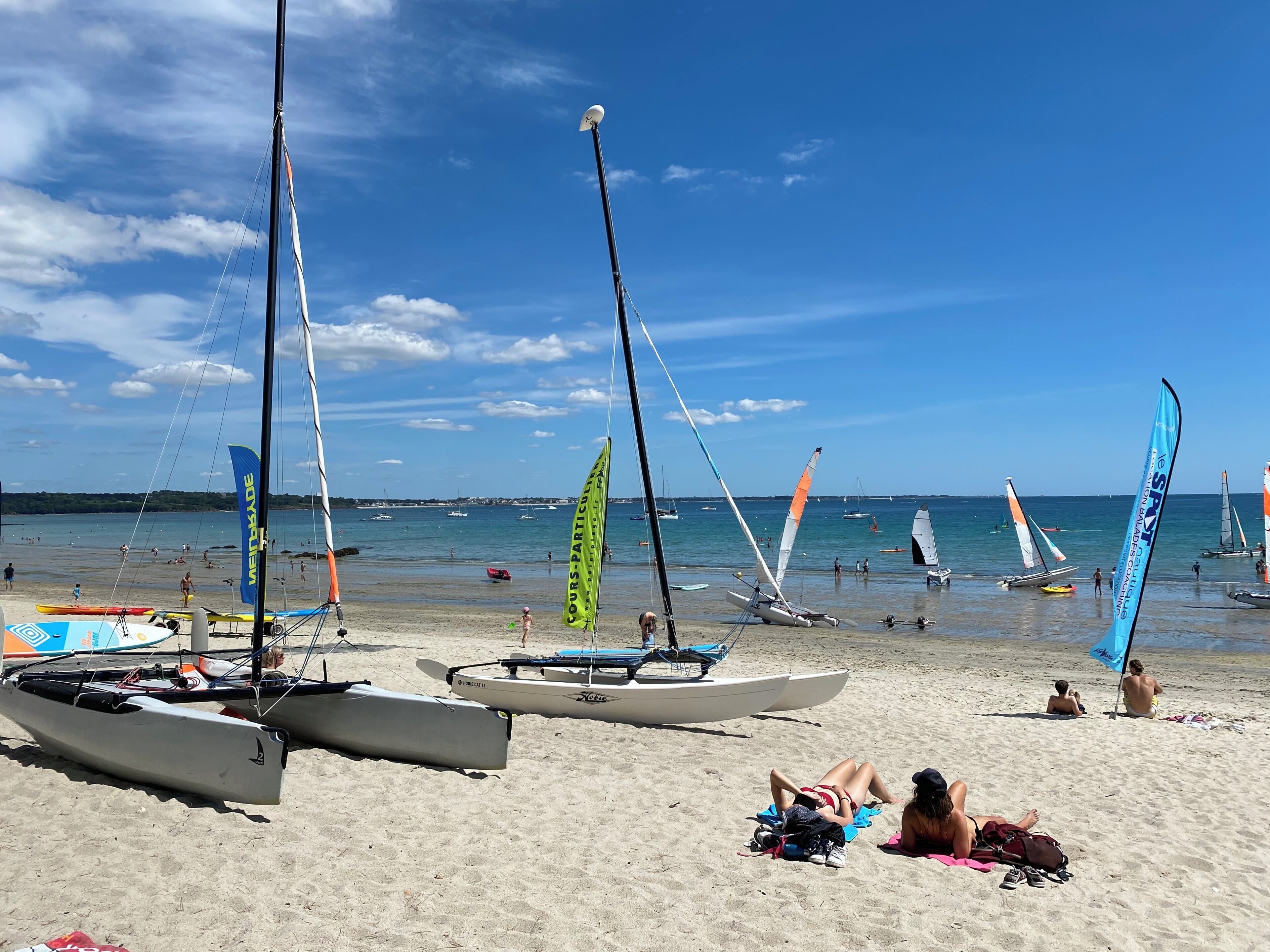 Uma praia com vários veleiros ancorados perto da costa, céu claro e pessoas relaxando na areia.