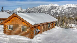 Uma cabana de toras com o telhado coberto de neve, rodeada por montanhas e árvores cobertas de neve.