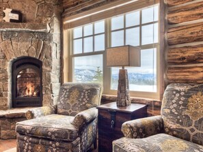 Ein rustikales Blockhaus-Interieur mit Kamin aus Stein, gemusterten Sesseln, einem Holznachttisch und einem großen Fenster mit Blick auf schneebedeckte Berge.
