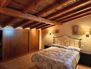 Une chambre à coucher avec un plafond en bois, un lit recouvert d’une literie à motifs floraux, deux lampes de chevet et une armoire.