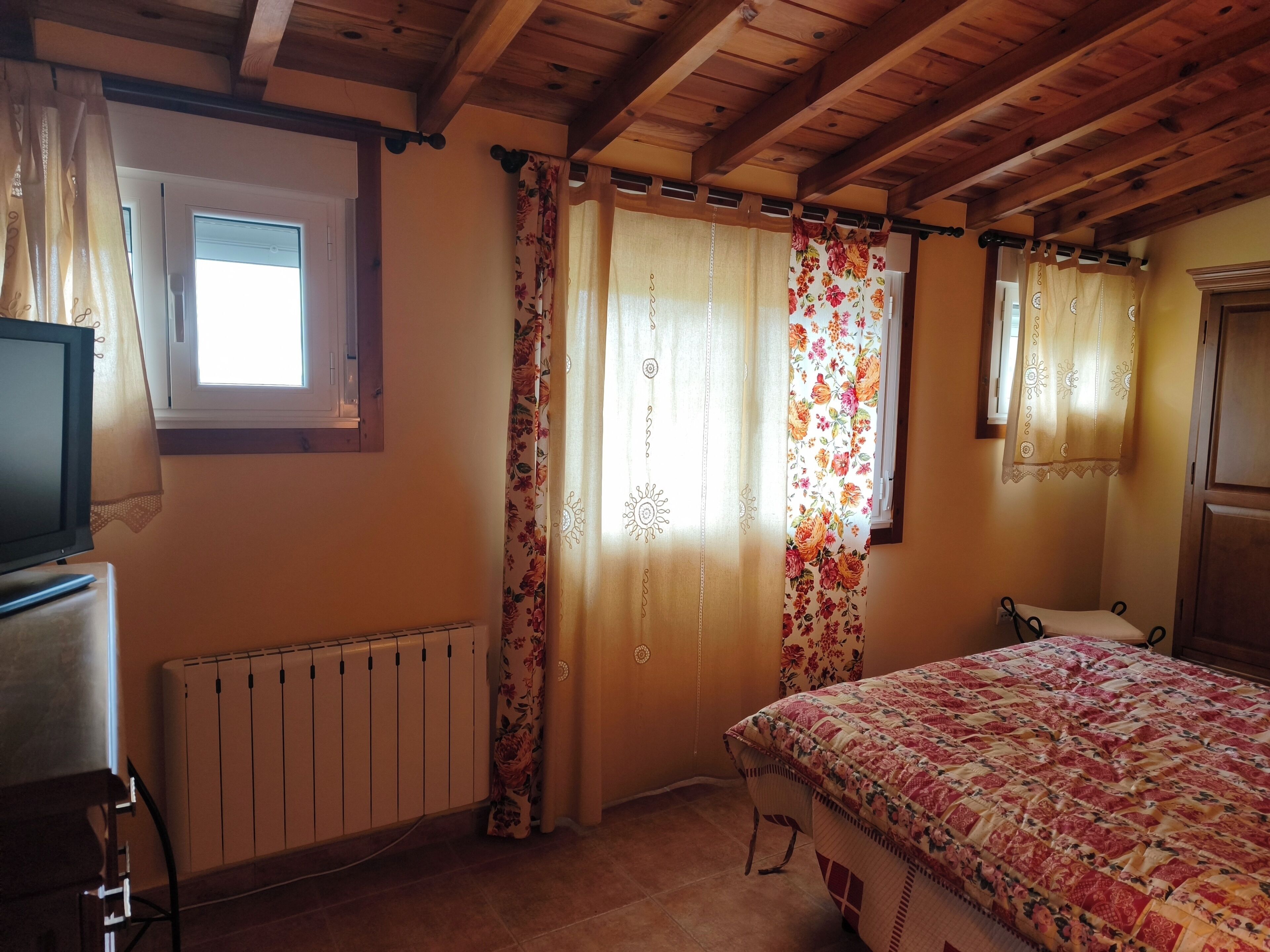 Un dormitorio con techo de madera, una cama con colcha estampada, un radiador y cortinas con motivos florales.