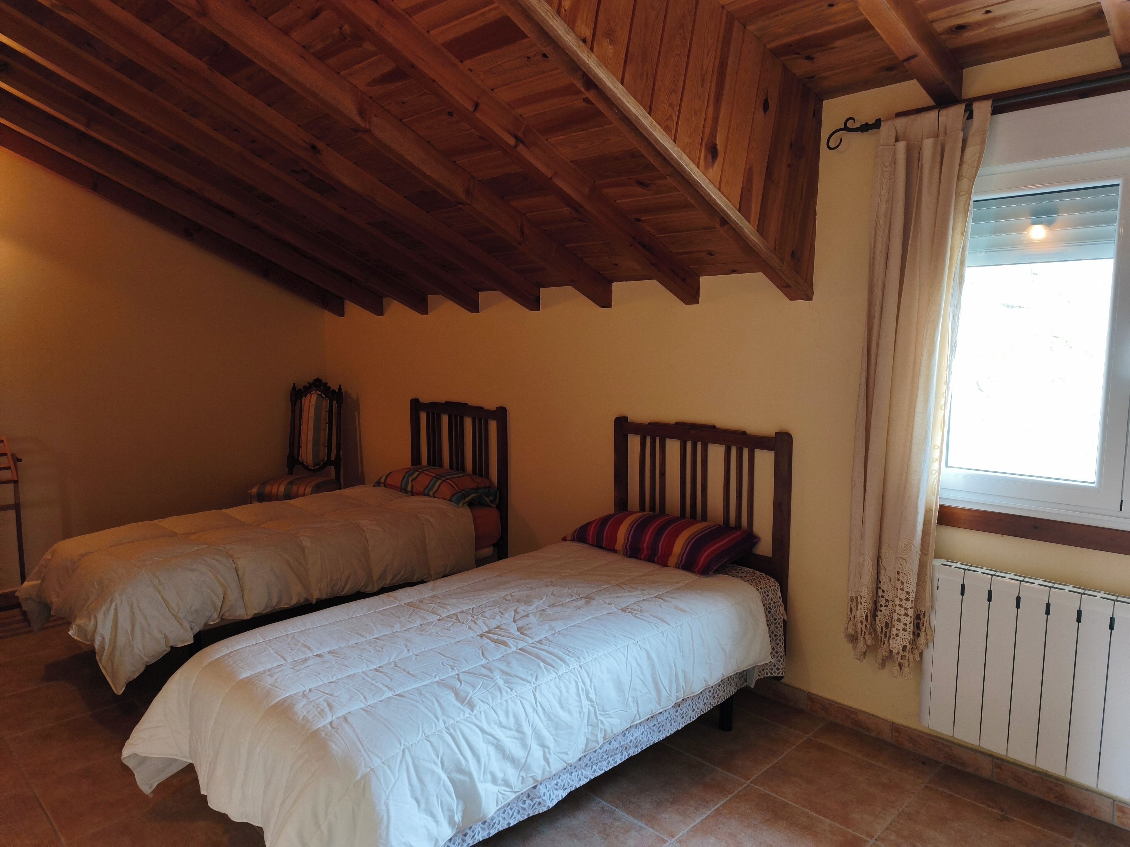 A bedroom with two beds, wooden beams on the ceiling, a window with curtains, and a radiator.