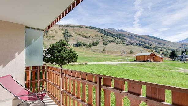 Un balcón con barandilla de madera, una silla roja y vistas a un campo verde y colinas.