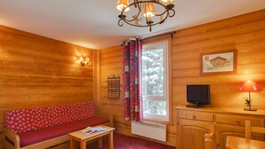 Interior de una cabaña de madera con sofá, mesa, televisor y una ventana con cortinas.