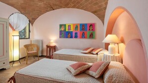 A bedroom with two beds, a wicker chair, a radiator, and a colorful artwork on the wall. - Oliveta Passero in Gavignano (Poggibonsi)