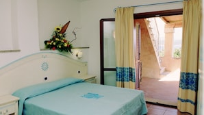 Un dormitorio con cama, mesita de noche, lámpara de pared, arreglo floral y vistas al balcón exterior.