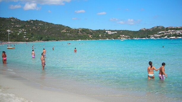 Una playa con agua turquesa cristalina, gente nadando y paseando por la orilla, y edificios al fondo.