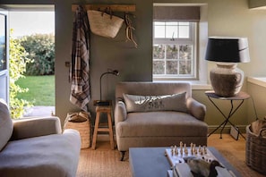 A cozy living room with a sofa, a small table with a lamp, a chessboard, and a window with a view of greenery. - Escape to a luxury 2 bdrm country cottage and ocean views (Kirkcaldy)
