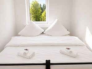 Ett litet, vitt rum med en enkel säng, ett fönster, vita sängkläder och två handdukar med ett lila blommönster.