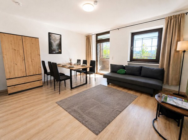 Ett modernt vardagsrum med en soffa, ett matbord med stolar, en trägarderob och ett stort fönster med gardiner.
